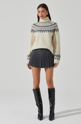 A woman is wearing an oversized, cream-colored turtleneck sweater with a gray and white geometric pattern around the yoke and sleeves. She pairs it with a high-waisted, dark gray pleated mini skirt. She completes the outfit with black, knee-high boots with a shiny finish.
